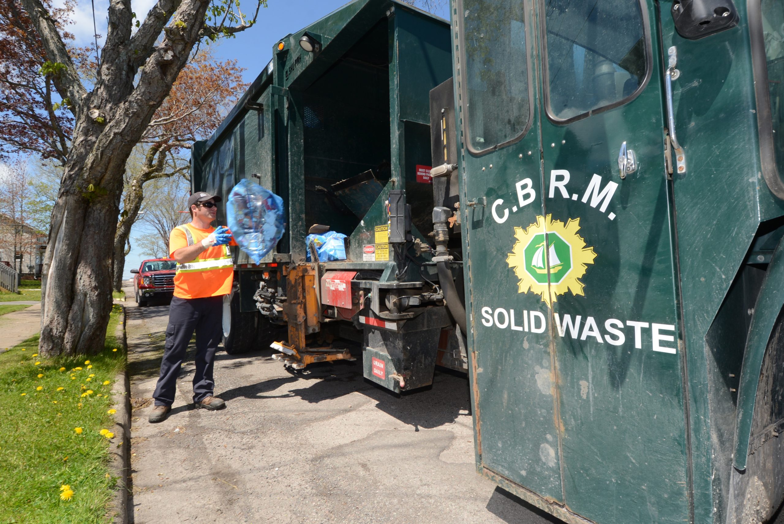 A waste collection worker wearing high-visibility safety gear places a blue recycling bag into a green CBRM Solid Waste truck.